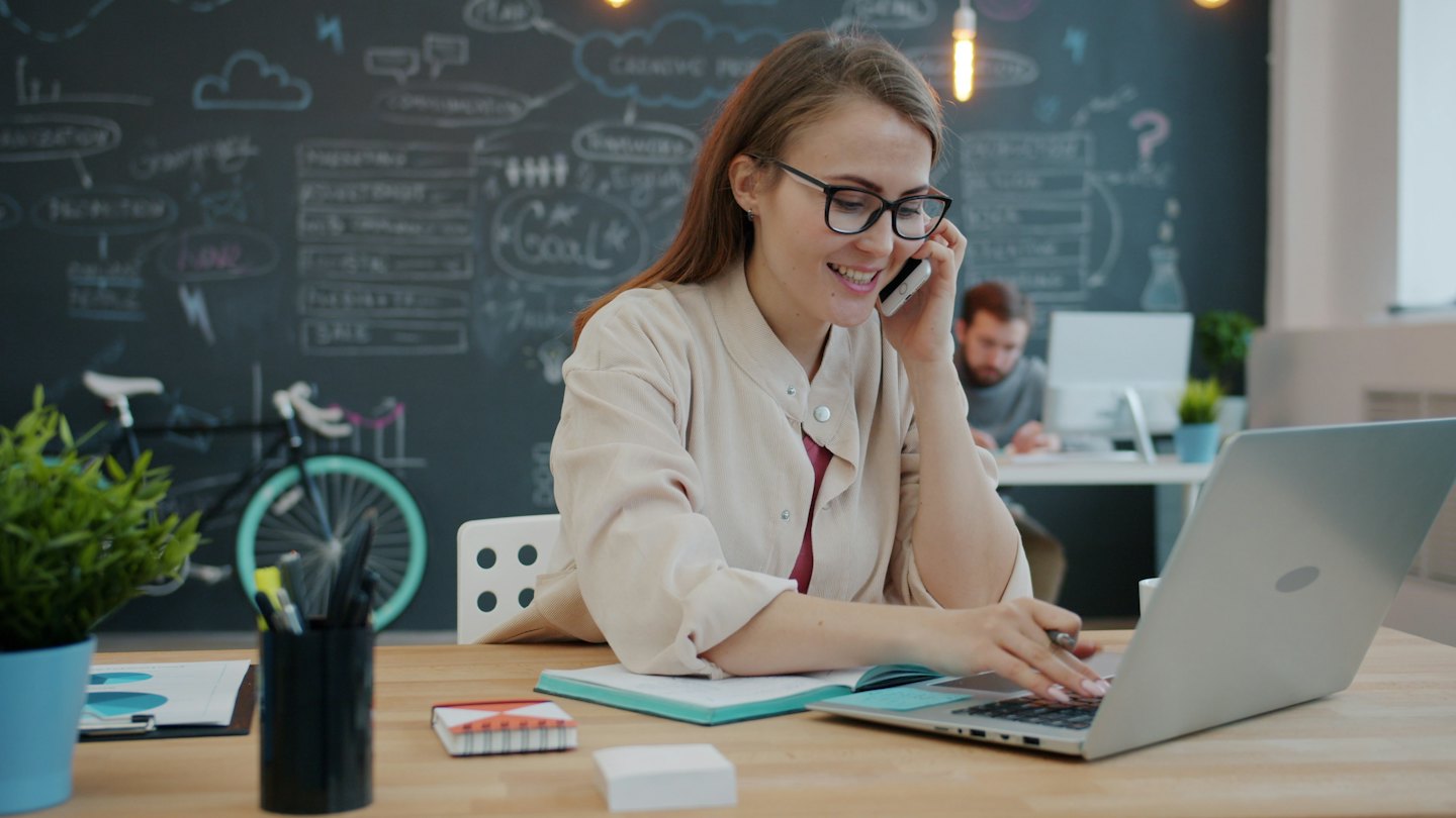 Woman smiling while talking on phone and typing on laptop at desk.