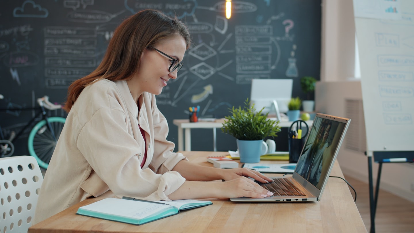 Woman with glasses smiles while typing on a laptop at a desk.